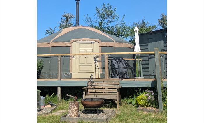 In front of the deck at the yurt, wood bench with wood framed gravel square containing Kadai fire pit with grill and cast iron skillet on the tripod a